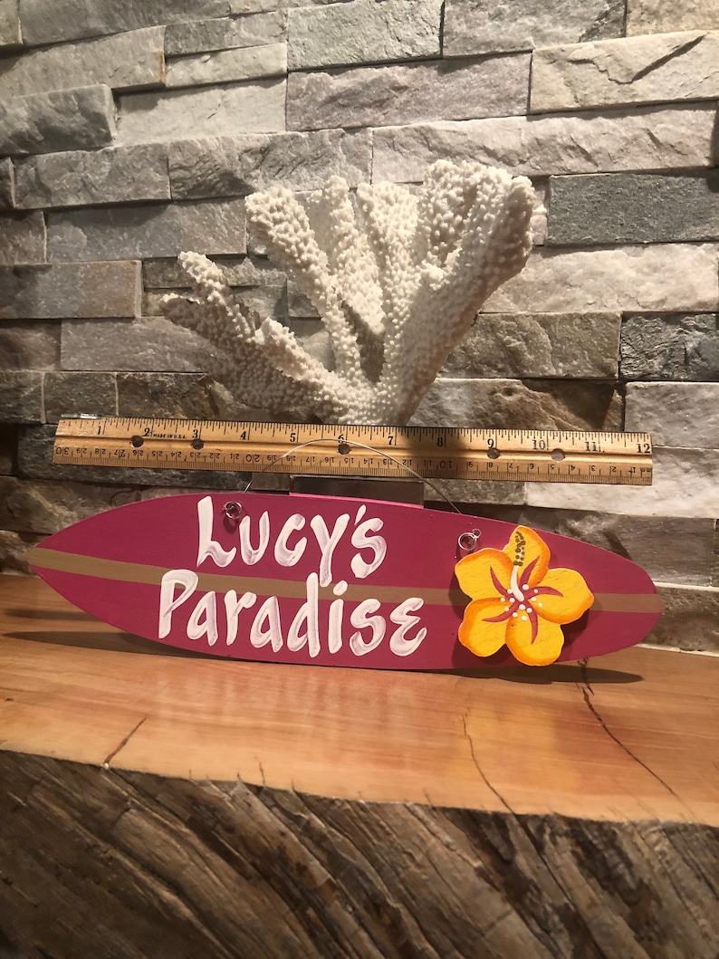 May include: A pink surfboard-shaped wooden sign with the text "Lucy's Paradise" in white paint and a yellow hibiscus flower painted on the right side.