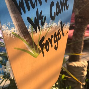 May include: A wooden sign with a blue and tan background. The sign has a painted image of a palm tree and white sand. The text on the sign reads "With Friends We Can't Forget..."