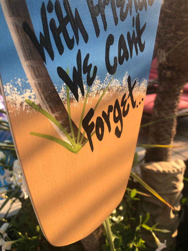 May include: A wooden sign with a blue and tan background. The sign has a painted image of a palm tree and white sand. The text on the sign reads "With Friends We Can't Forget..."