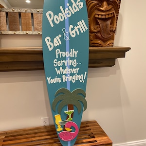May include: A blue surfboard sign with the text "Poolside Bar & Grill" and "Proudly Serving... Whatever You're Bringing!"  The sign has a pink flamingo and palm trees with drinks on it.