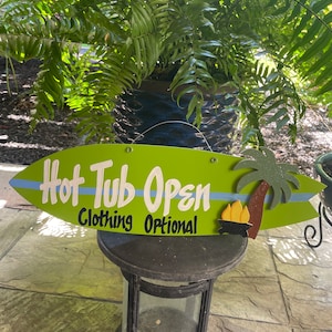 May include: A green surfboard-shaped sign with the text "Hot Tub Open" and "Clothing Optional" in white lettering. The sign also features a palm tree and a campfire graphic.
