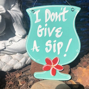 May include: A turquoise blue wooden sign shaped like a cocktail glass with the text "I Don't Give A Sip!" in white paint. A red flower with a yellow center is painted on the bottom of the glass.