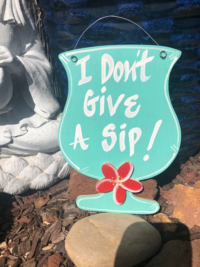 May include: A turquoise blue wooden sign shaped like a cocktail glass with the text "I Don't Give A Sip!" in white paint. A red flower with a yellow center is painted on the bottom of the glass.