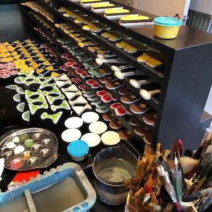 May include: A close-up of a work table with a variety of colorful ceramic tiles, paint, brushes, and other supplies. The tiles are arranged on shelves and are ready to be painted.