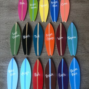 Op de afbeelding: Twintig miniatuur surfplank ornamenten geschilderd in verschillende kleuren, waaronder roze, groen, geel, blauw, koraal, zwart, teal, oranje, wijn, turquoise, babyblauw, aqua, rood, bruin, paars en oceaanblauw. Elke surfplank heeft een witte streep in het midden en een kleurnaam erop geschreven.