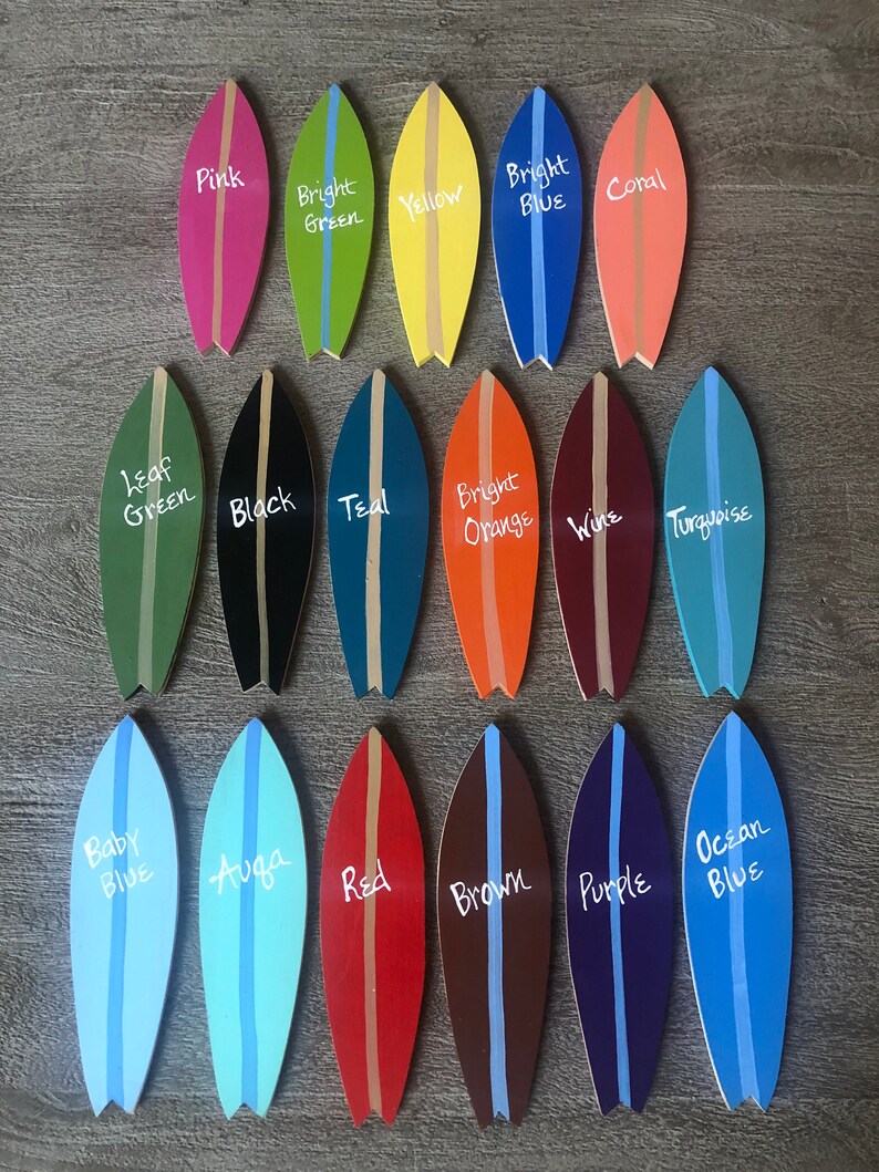 Op de afbeelding: Twintig miniatuur surfplank ornamenten geschilderd in verschillende kleuren, waaronder roze, groen, geel, blauw, koraal, zwart, teal, oranje, wijn, turquoise, babyblauw, aqua, rood, bruin, paars en oceaanblauw. Elke surfplank heeft een witte streep in het midden en een kleurnaam erop geschreven.