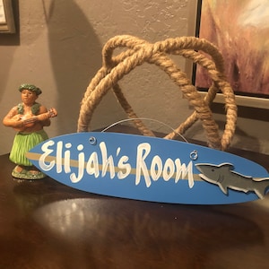 May include: Blue surfboard-shaped wooden sign with the text "Elijah's Room" in white paint and a gray shark illustration. The sign is hanging from a rope.
