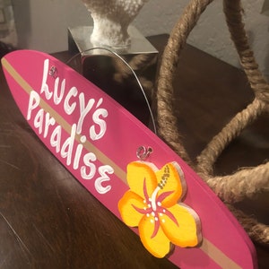 May include: Pink wooden surfboard sign with the text "Lucy's Paradise" in white paint. A yellow hibiscus flower is painted on the sign.