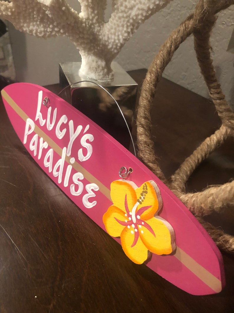 May include: Pink wooden surfboard sign with the text "Lucy's Paradise" in white paint. A yellow hibiscus flower is painted on the sign.