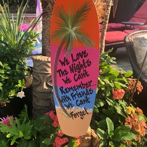 May include: A colorful wooden surfboard with a painted palm tree and the text "We Love The Nights We Can't Remember... With Friends We Can't Forget..."