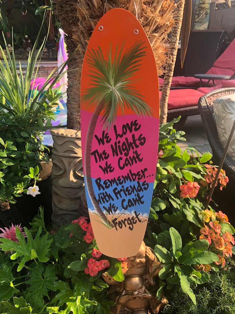 May include: A colorful wooden surfboard with a painted palm tree and the text "We Love The Nights We Can't Remember... With Friends We Can't Forget..."