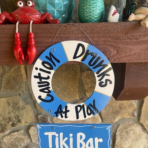 May include: A blue and white life preserver sign with the text "Caution Drunks At Play" hanging from a wooden shelf. Below the sign is a blue sign with the text "Tiki Bar" in white.