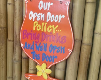 May include: A wooden sign shaped like a cocktail glass with a yellow flower at the bottom. The sign is orange and has the text "Our Open Door Policy... Bring Drinks And We'll Open The Door" written in colorful letters.