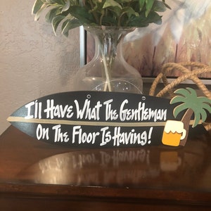 May include: Black surfboard-shaped wooden sign with white lettering that reads "I'll Have What The Gentleman On The Floor Is Having!". A palm tree and beer mug are on the right side. The sign is a decorative item.