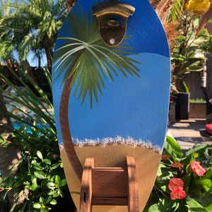 May include: A wooden surfboard-shaped bottle opener with a palm tree and beach scene painted on it. The bottle opener has a metal bottle opener attached to the top and a wooden holder on the bottom.