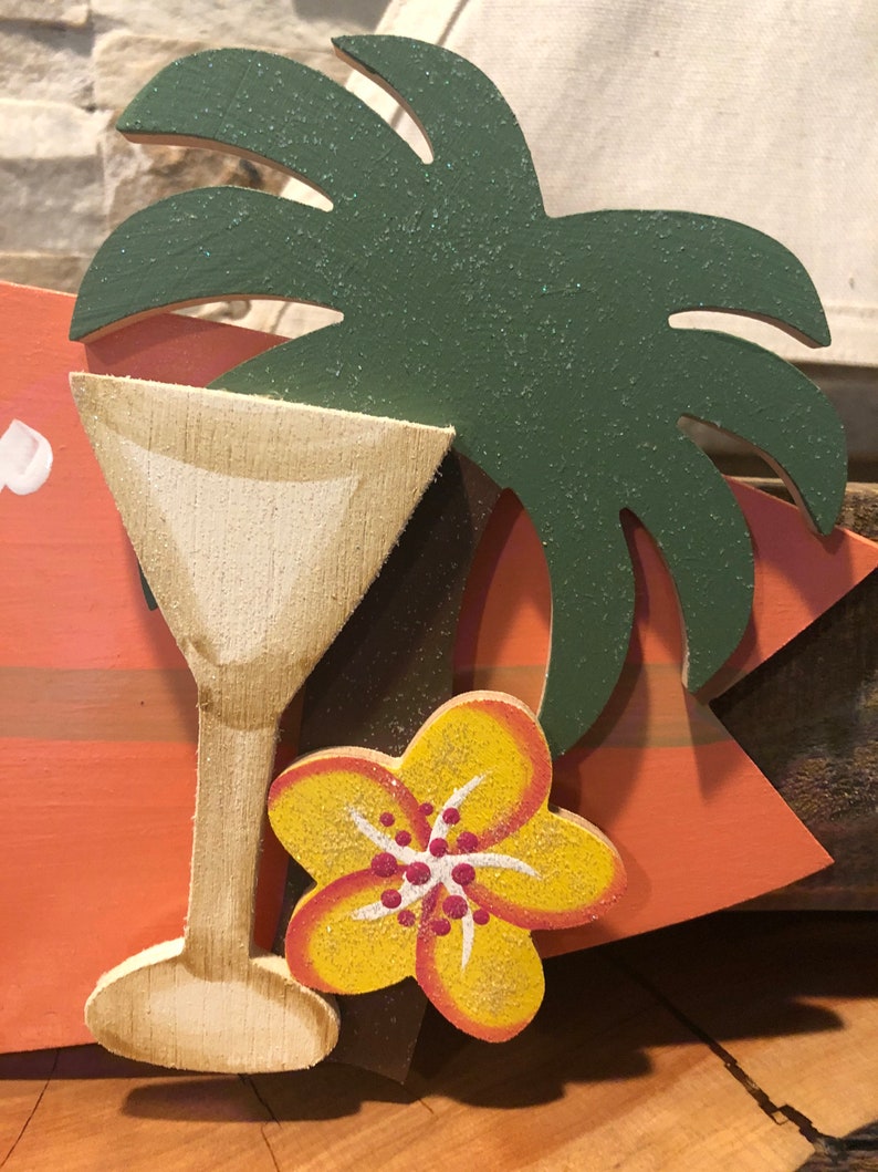 Op de afbeelding: Een houten bord met een geschilderd beeld van een tropisch drankje, een gele bloem en een groene palmboom. Het bord is geschilderd in tinten oranje, geel, groen en bruin.
