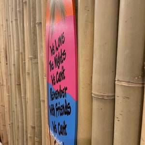 May include: A pink and blue surfboard with a palm tree and the text "We love the nights we can't remember with friends we can't forget..." hanging on a bamboo wall.