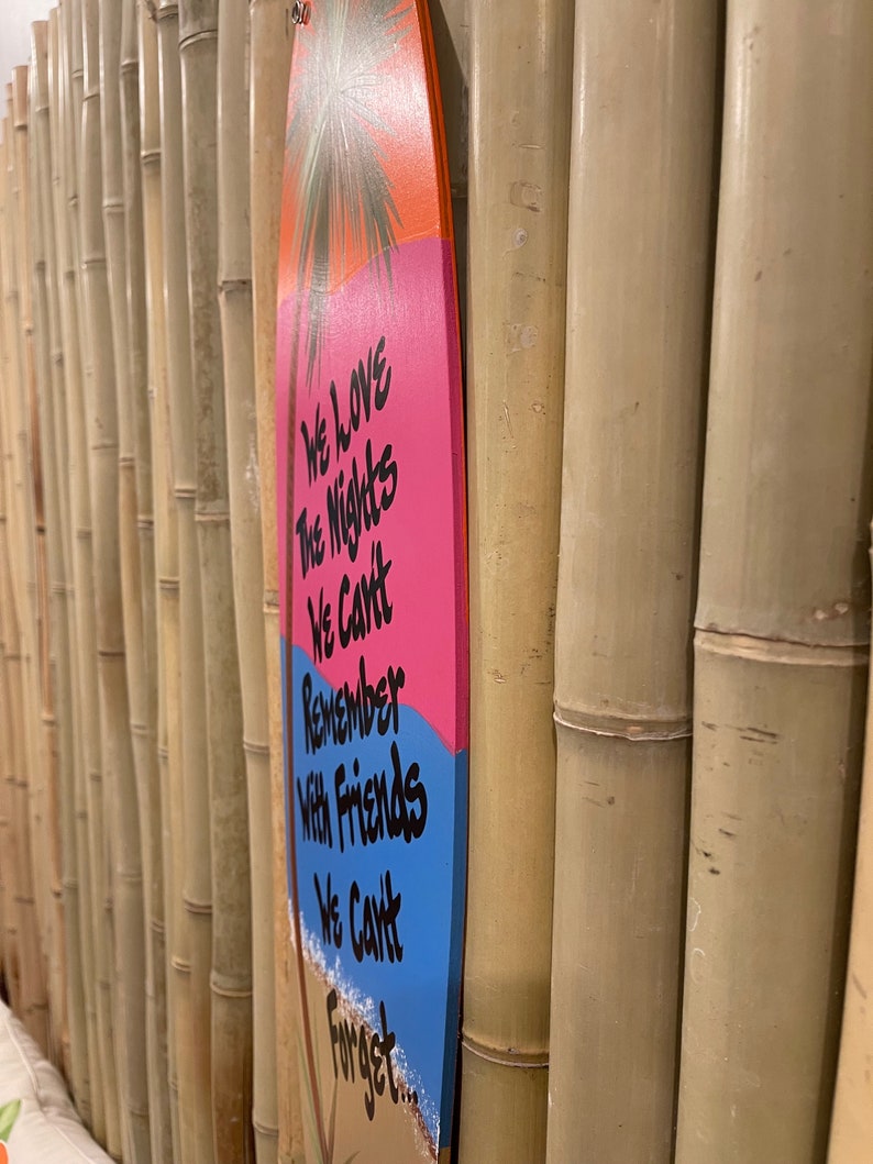 May include: A pink and blue surfboard with a palm tree and the text "We love the nights we can't remember with friends we can't forget..." hanging on a bamboo wall.
