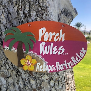 May include: A wooden oval sign with the text "Porch Rules. Relax*Party*Relax" in white paint. The sign is painted in orange, pink, and brown with a green palm tree and a yellow flower.