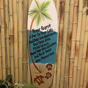 May include: A wooden surfboard-shaped sign with a blue and green ocean scene and a palm tree. The sign has the text "Never Regret A Day In Your Life... Good Days Give Happiness. Bad Days Give Experience. Worst Days Give Lessons. And Best Days Give Memories."