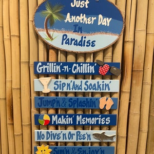 May include: A set of wooden signs with blue and white paint and various beach-themed illustrations. The top sign reads "Just Another Day In Paradise". The other signs read "Grillin'-n-Chillin', Sip n'And Soakin', Jump'n & Splashin', Makin' Memories, No Dive'n Or Pee'n, Sun'n & Enjor'n".