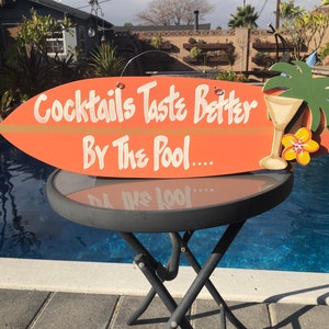 Op de afbeelding: Een oranje surfplank-vormig houten bord met de tekst "Cocktails Taste Better By The Pool..." in witte verf. Aan de rechterkant van het bord is een groene palmboom met een gele bloem en een glas vloeistof geschilderd.
