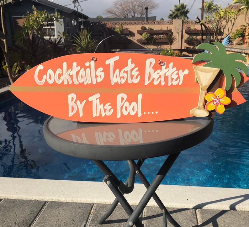 Op de afbeelding: Een oranje surfplank-vormig houten bord met de tekst "Cocktails Taste Better By The Pool..." in witte verf. Aan de rechterkant van het bord is een groene palmboom met een gele bloem en een glas vloeistof geschilderd.