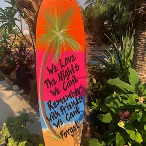 May include: A wooden surfboard-shaped sign with a painted palm tree and the text "We love the nights we can't remember... with friends we can't forget..."