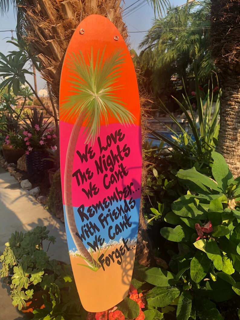 May include: A wooden surfboard-shaped sign with a painted palm tree and the text "We love the nights we can't remember... with friends we can't forget..."