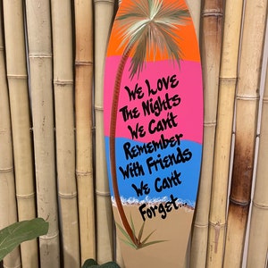 May include: A wooden surfboard-shaped sign with a painted palm tree, ocean waves, and the text "We love the nights we can't remember with friends we can't forget."