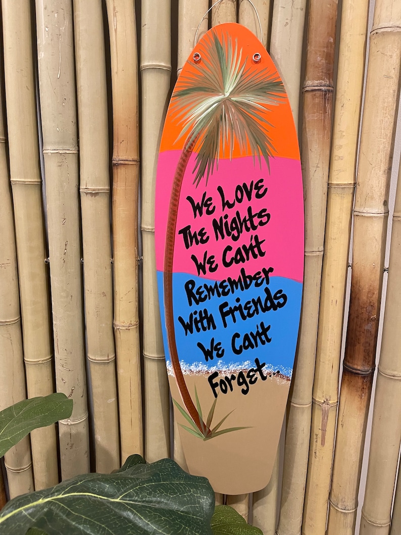 May include: A wooden surfboard-shaped sign with a painted palm tree, ocean waves, and the text "We love the nights we can't remember with friends we can't forget."