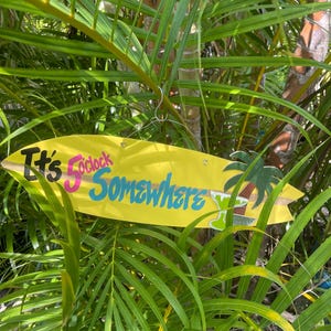 May include: A yellow surfboard-shaped sign with the text "It's 5 o'clock Somewhere" in black, pink, and blue. A palm tree and cocktail graphic are on the right. The sign is hanging among green palm leaves.