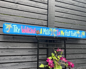 Puede incluir: Un letrero azul y blanco con el texto "Try Walking a Mile In My Flip Flops" con palmeras y chanclas a cada lado del texto.