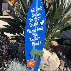 Puede incluir: Un cartel azul en forma de tabla de surf con una palmera blanca y una flor de hibisco roja. El cartel dice "We Don't Swim In Your Toilet... Please Don't Pee... In Our Pool!"
