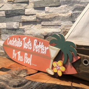 Op de afbeelding: Een houten surfboard-bord met de tekst "Cocktails Taste Better By The Pool..." in wit geschilderd. Het bord is versierd met een groene palmboom, een gele bloem en een martini-glas.