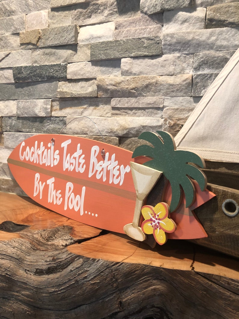 Op de afbeelding: Een houten surfboard-bord met de tekst "Cocktails Taste Better By The Pool..." in wit geschilderd. Het bord is versierd met een groene palmboom, een gele bloem en een martini-glas.