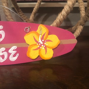 May include: Pink wooden surfboard with a yellow hibiscus flower painted on it. The surfboard has the word "House" written on it in white paint.