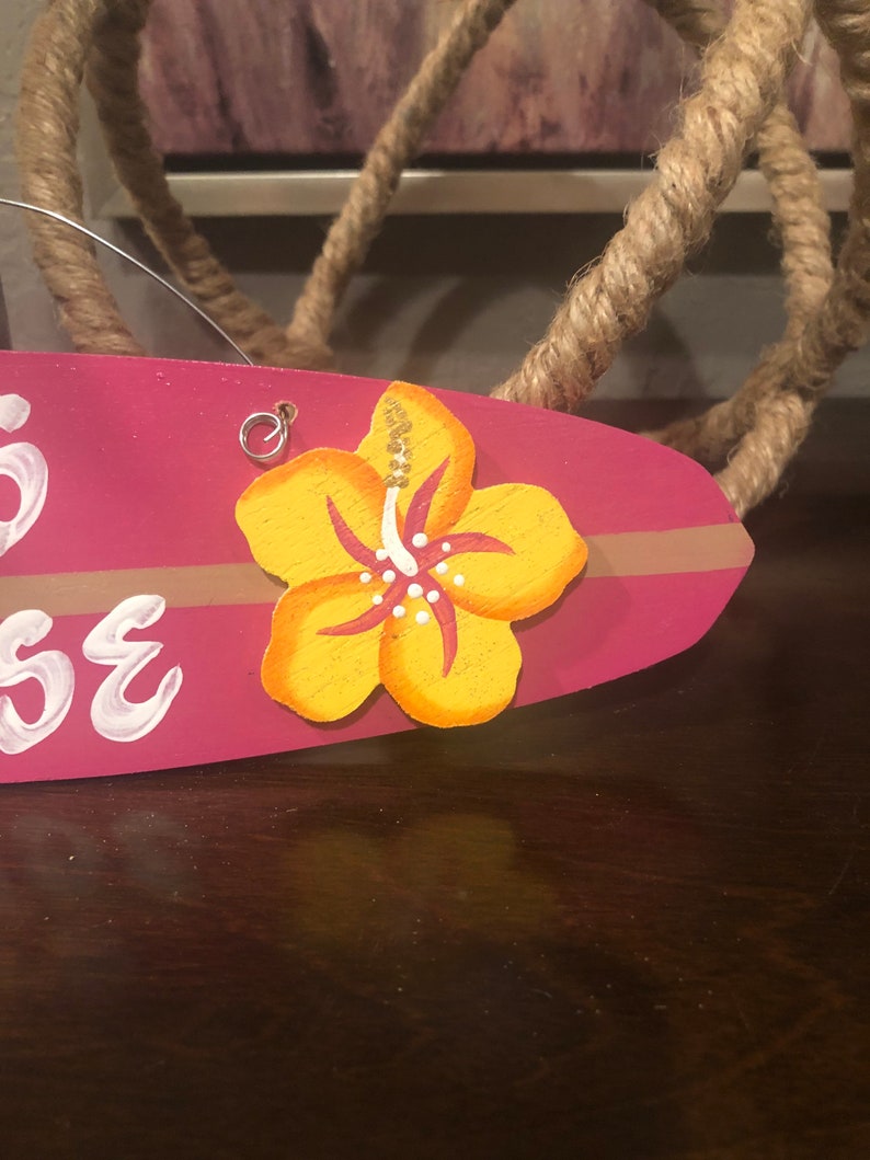 May include: Pink wooden surfboard with a yellow hibiscus flower painted on it. The surfboard has the word "House" written on it in white paint.