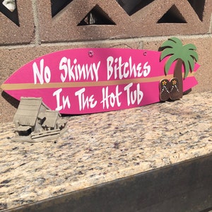 Puede incluir: Un cartel de surf rosa con el texto "No Skinny Bitches In The Hot Tub" en letras blancas. Un pequeño palmeral con chanclas marrones está pintado en el lado derecho del cartel.