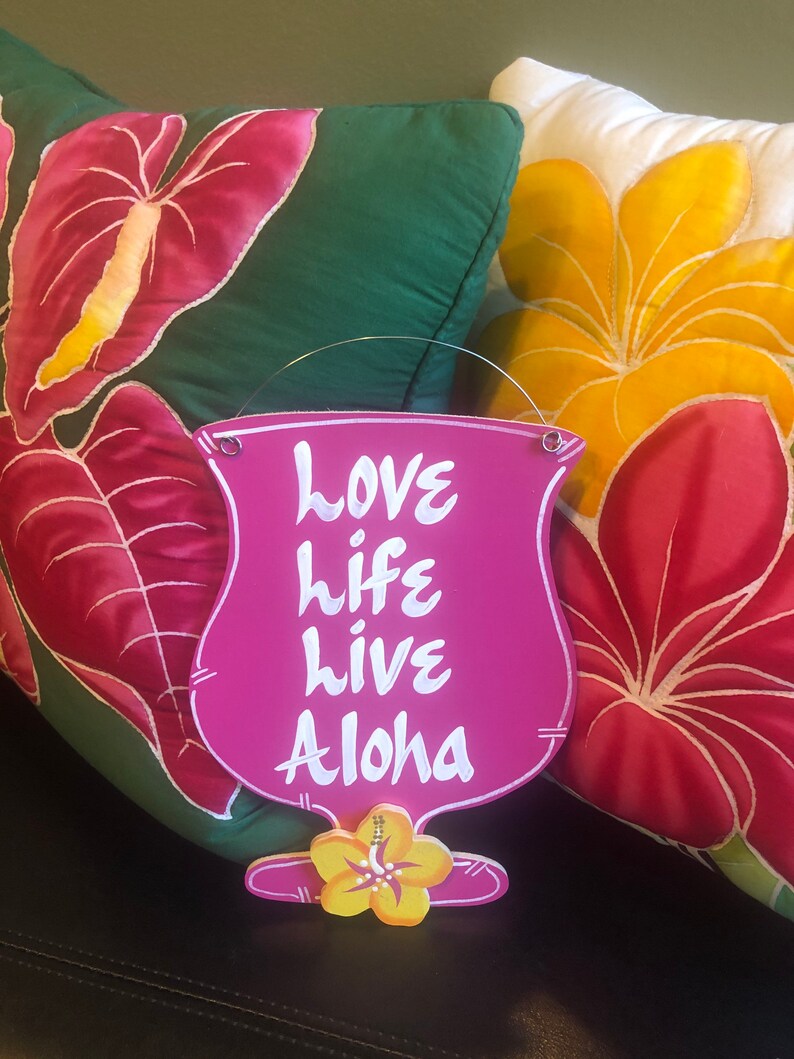 May include: Pink wooden sign shaped like a cocktail glass with the words "love life live Aloha" written in white. The sign is hanging from two small metal rings.