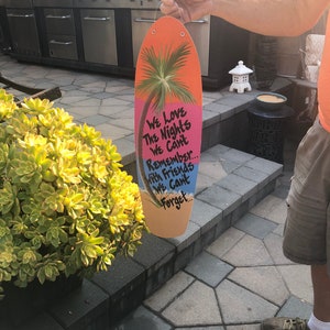 May include: A pink and orange surfboard-shaped sign with a palm tree and the text "We Love The Nights We Can't Remember... With Friends We Can't Forget."