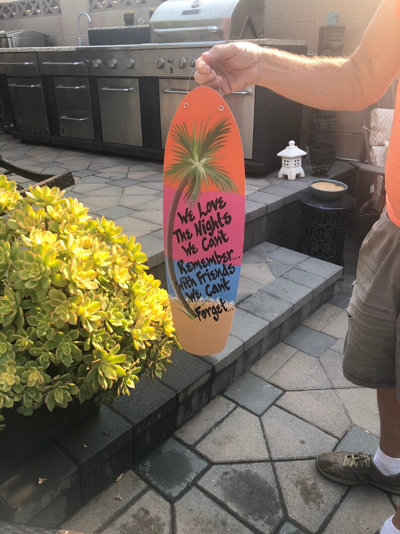 May include: A pink and orange surfboard-shaped sign with a palm tree and the text "We Love The Nights We Can't Remember... With Friends We Can't Forget."