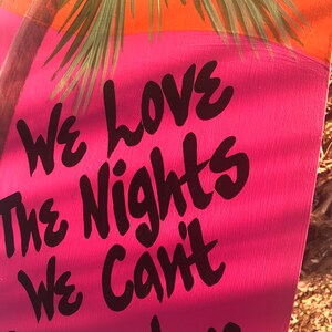 May include: A pink and blue sign with black text that reads "We love the nights we can't remember..." The sign is painted with a palm tree in the background.