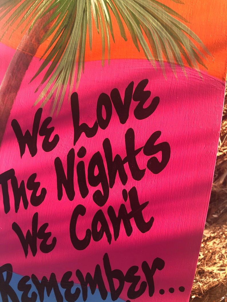 May include: A pink and blue sign with black text that reads "We love the nights we can't remember..." The sign is painted with a palm tree in the background.
