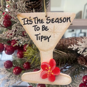 May include: A wooden ornament shaped like a martini glass, with the text "It's The Season To Be Tipsy". The ornament has a red flower attached and is hanging on a Christmas tree with red berries.