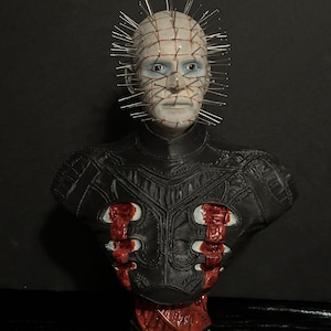May include: A detailed bust of a character with a pale face, blue eyes, and a grid-like pattern on the head with numerous metal pins. The figure wears a black leather-like garment with red accents and sits atop a decorative base.