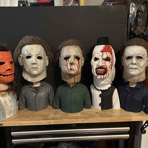 May include: Five horror movie character busts are displayed on a wooden shelf. The masks include a pumpkin, Michael Myers, and a clown with blood. The busts are on a wooden shelf.