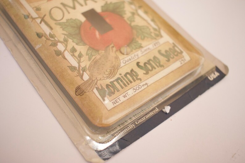 May include: A vintage-style light switch cover with a brown background and a colorful illustration of a tomato plant, a bird, and the text "Morning Song Seed, Quality Seed, Net Wt. 300mg, 254,  Gauranteed, USA".