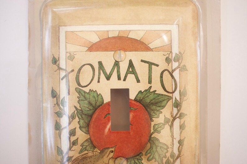 May include: A light switch cover with a vintage illustration of a tomato plant and the word "TOMATO" in a decorative font. The switch cover is made of clear plastic and has a white background.