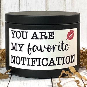 Può includere: Una candela nera con un'etichetta bianca con la scritta "YOU ARE MY favorite NOTIFICATION" e un'impronta di labbra rossa. La candela ha un coperchio nero ed è circondata da carta marrone.